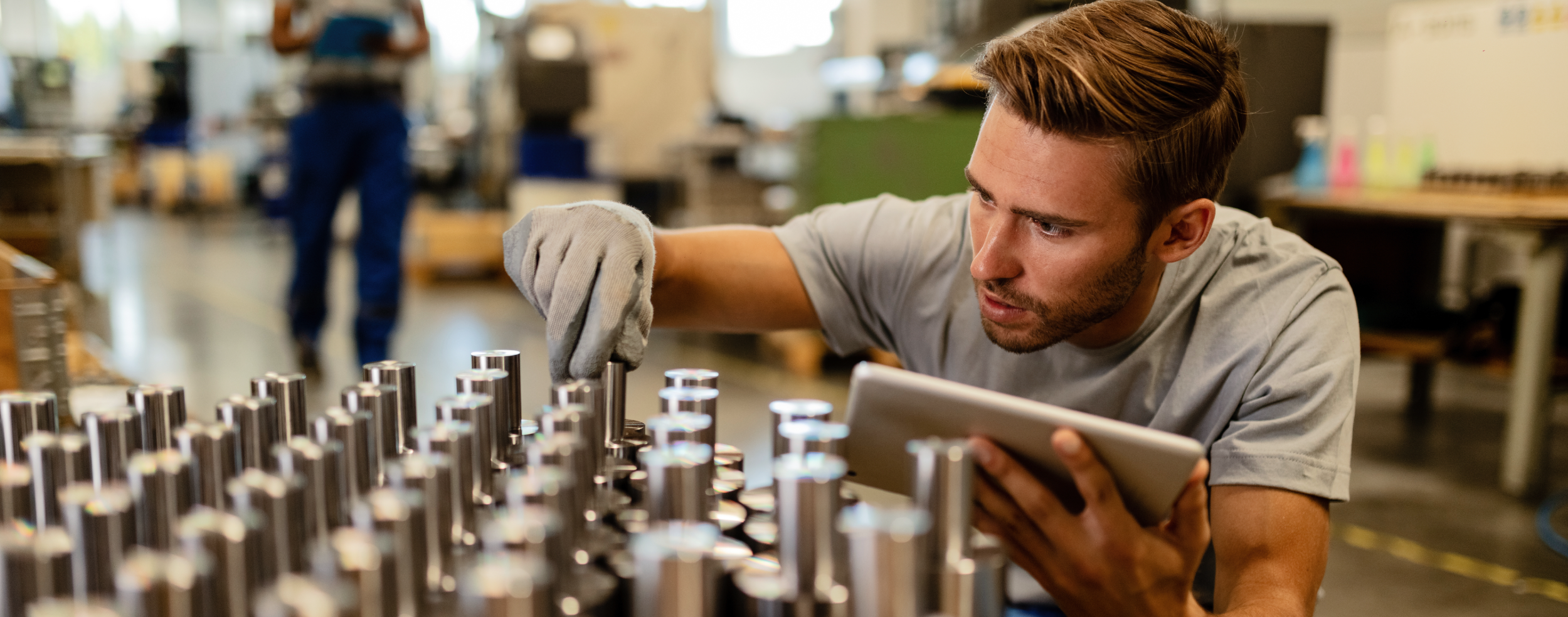 Man wearing a glove inspects metal cylindrical components on a production line, holding a tablet.