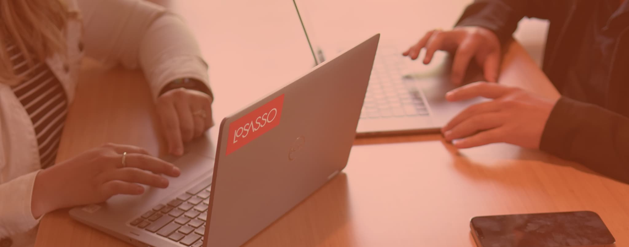 Two people working on laptops at a wooden table, collaborating in a casual office setting.